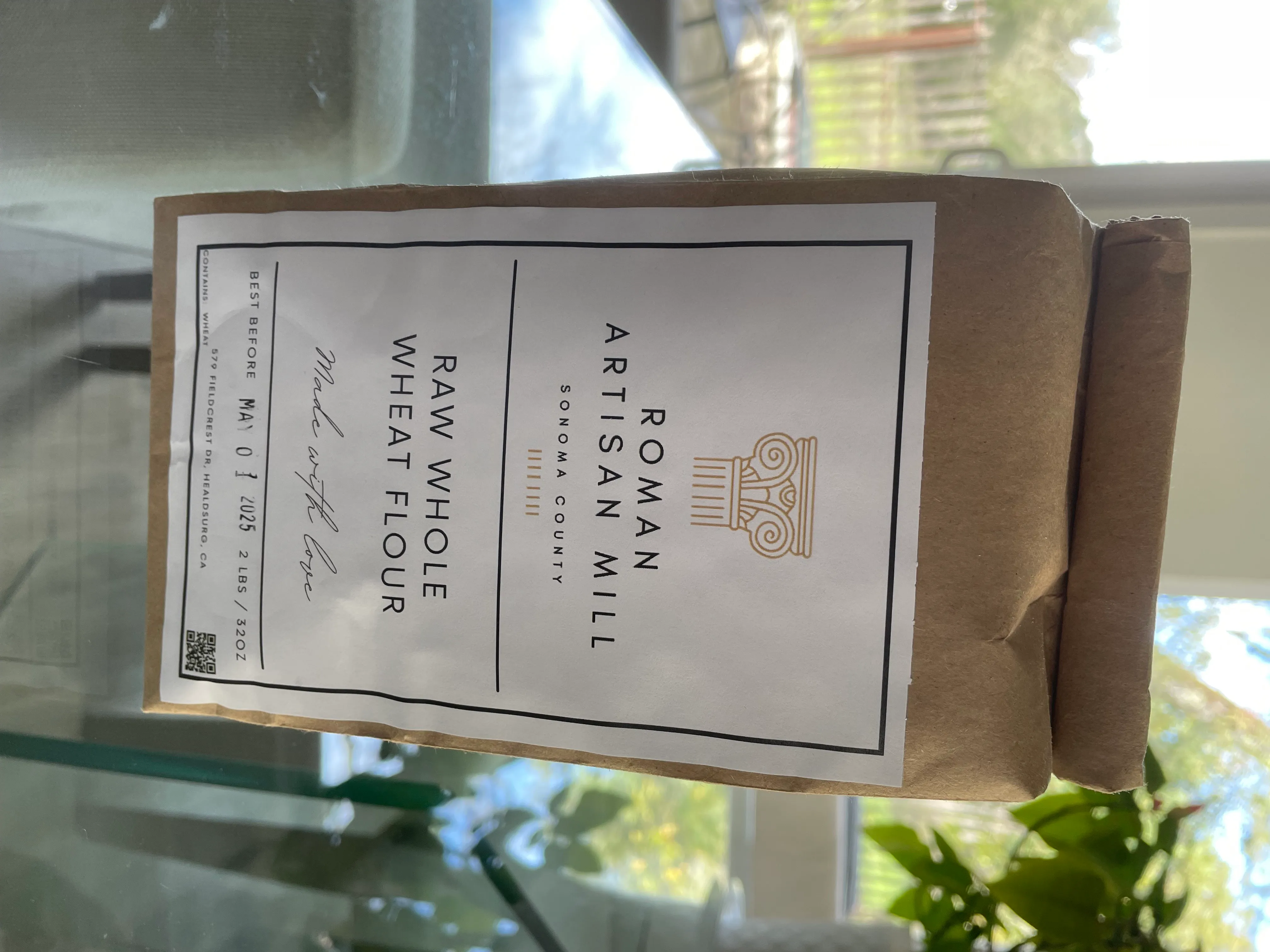 The Roman Artisan Mill kraft bag with its white label, gold column mark, and letter-spaced caps reading Raw Whole Wheat Flour